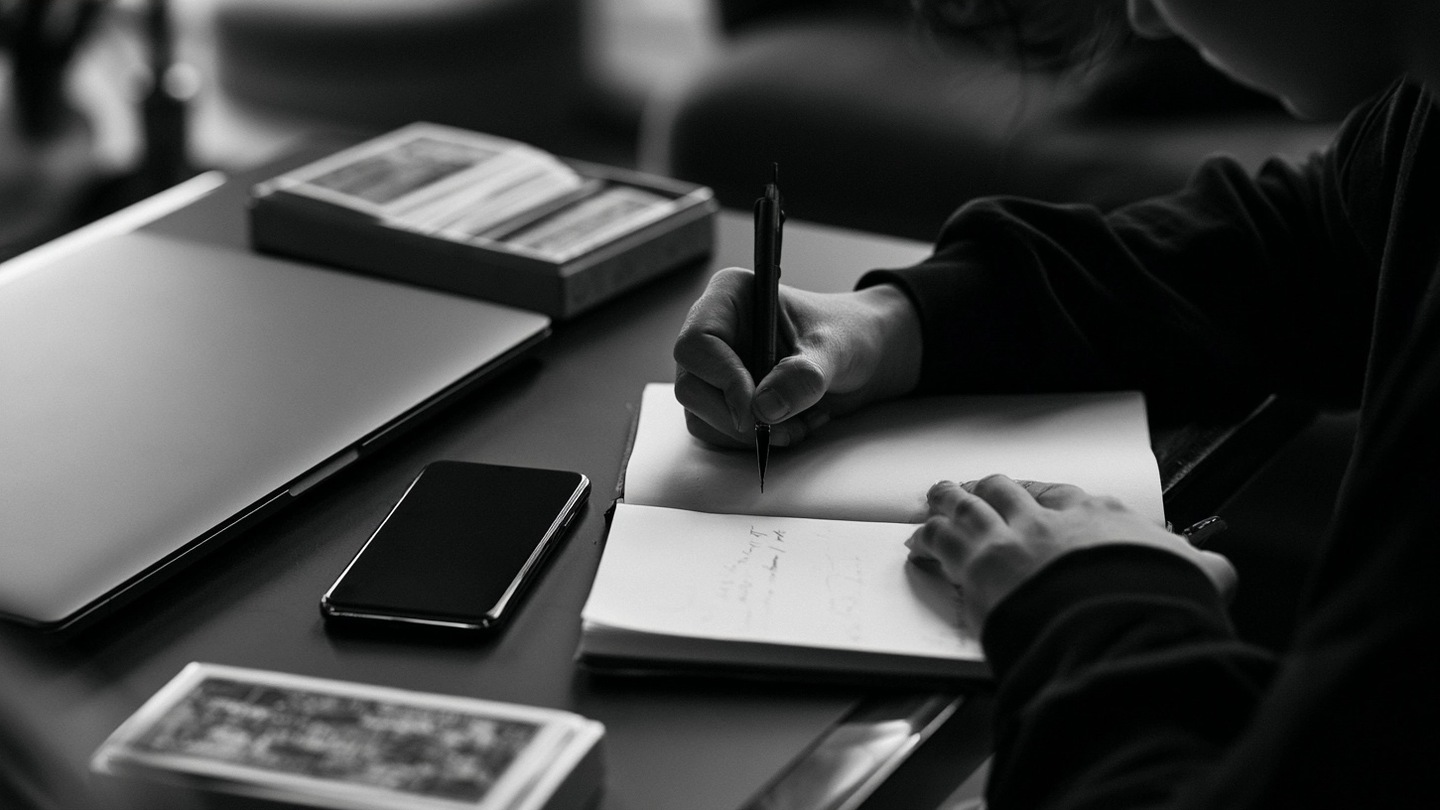 Une personne pose son téléphone et note des faits près d’un ordinateur fermé et d’un tarot rangé.